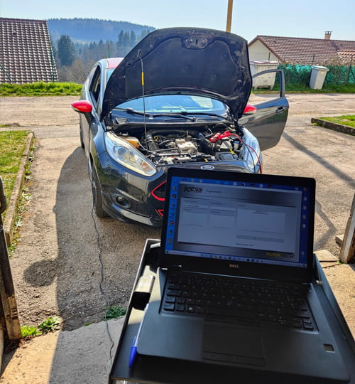 Une Ford grise avec le capot ouvert, en cours de reprogrammation moteur, un ordinateur portable affichant les paramètres du moteur est posé à proximité.